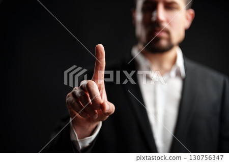 Man in a suit holds one finger up against a dark background to convey a message or emphasize a point during a serious discussion 130756347