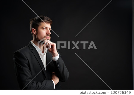 Businessman in formal suit pondering ideas against dark background in a professional setting Businessman in formal suit pondering ideas against dark background in a professional setting 130756349