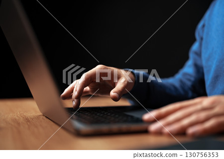 Person working on a laptop at a wooden desk during evening hours in a dimly lit room Person working on a laptop at a wooden desk during evening hours in a dimly lit room 130756353