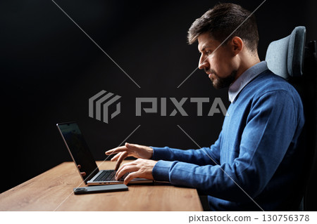Focused individual working on a laptop at a wooden desk in a dimly lit room during evening hours 130756378