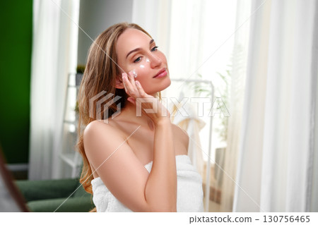 Young woman applying facial cream in a bright, cozy indoor setting during the day 130756465