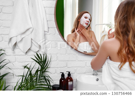 Woman applying face mask while looking at mirror in a bright bathroom with white brick walls and plants Woman applying face mask while looking at mirror in a bright bathroom with white brick walls and plants 130756466