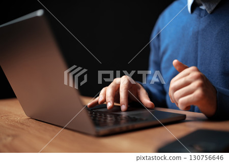 Focused individual working on a laptop at a wooden desk in a dimly lit room during evening hours Focused individual working on a laptop at a wooden desk in a dimly lit room during evening hours 130756646