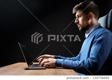 Focused man working on laptop in a dimly lit room during evening hours Focused man working on laptop in a dimly lit room during evening hours 130756660