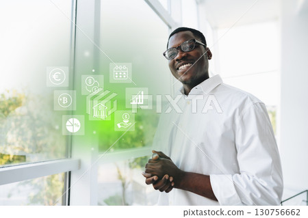 Professional man in a white shirt smiles confidently while standing by a large window with financial icons around him in an office setting Professional man in a white shirt smiles confidently while standing by a large window with financial icons around him in an office setting 130756662