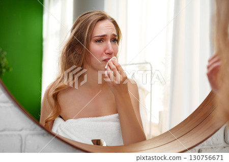 Woman removing makeup in front of mirror after long day, expressing emotions in a bright bathroom setting 130756671