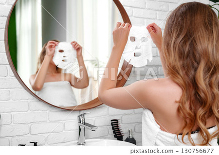 Woman applying facial mask in a bright bathroom while looking in the mirror 130756677
