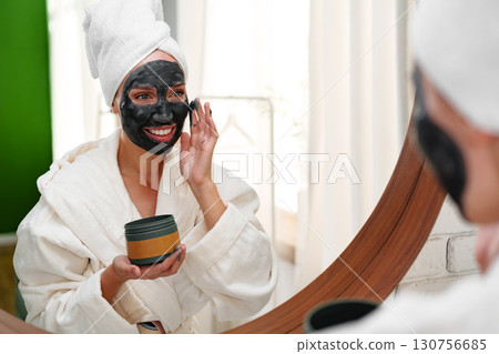 Woman applying face mask in bathrobe while smiling in front of mirror in bright room 130756685