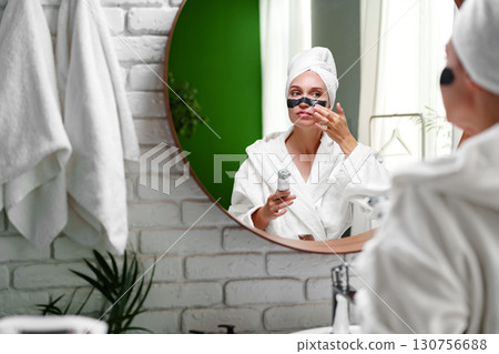 Woman enjoying a spa day at home while smiling and posing with facial mask in a modern bathroom Woman enjoying a spa day at home while smiling and posing with facial mask in a modern bathroom 130756688