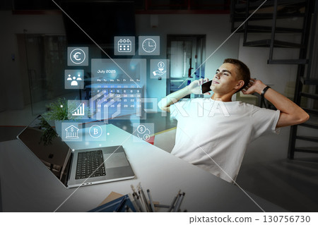Young man engages in remote work while using a laptop and phone in a futuristic office setting during the evening hours 130756730