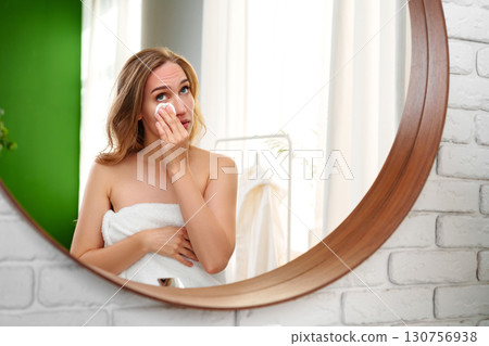 Woman applying skincare in a bright bathroom while looking in a round mirror Woman applying skincare in a bright bathroom while looking in a round mirror 130756938