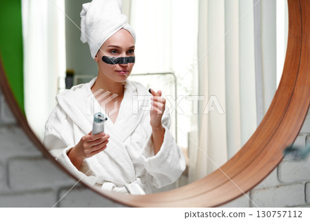 Woman enjoys skincare routine at home using facial roller in bathroom while wearing white bathrobe and towel Woman enjoys skincare routine at home using facial roller in bathroom while wearing white bathrobe and towel 130757112