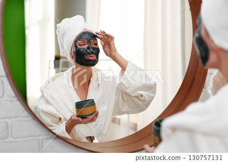 Woman in robe applying facial mask while looking in mirror during home spa time Woman in robe applying facial mask while looking in mirror during home spa time 130757131