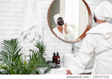 Woman applying face mask while enjoying self-care routine in bright bathroom decorated with plants 130757133