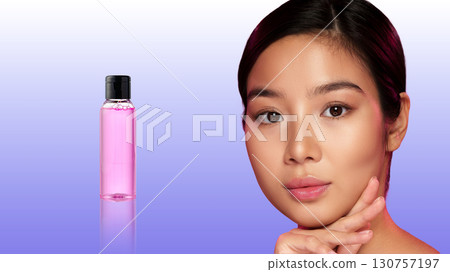 Close-up of a woman with glowing skin and a bottle of pink liquid against a gradient background showcasing beauty and skincare 130757197