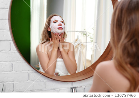 Woman applies facial mask while looking in mirror during self-care routine at home in bright, modern bathroom Woman applies facial mask while looking in mirror during self-care routine at home in bright, modern bathroom 130757466