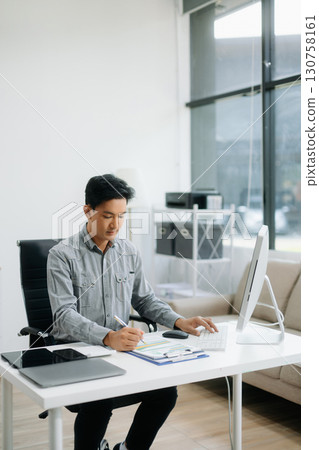 Confident businessman working on finance analysis with a tablet and laptop at office desk. 130758161