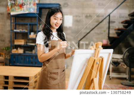 Cheerful female artist painting in a vibrant studio with brushes, a palette, and a blank canvas. 130758249