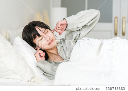 A young Asian woman covering her ears from noise while sleeping in bed (noise trouble, sleep disturbance, sleep disorder) 130758883