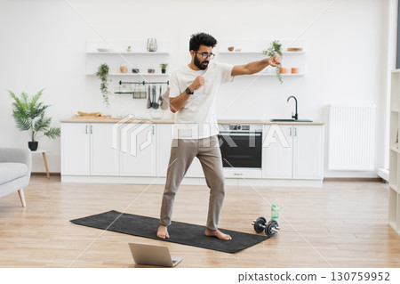 Middle-aged man doing fitness boxing exercises in bright kitchen, barefoot, using yoga mat. Middle-aged man doing fitness boxing exercises in bright kitchen, barefoot, using yoga mat. 130759952