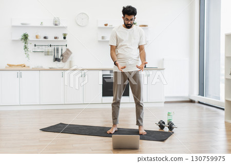 Young male adult performing exercises on mat in bright kitchen using online guidance. Young male adult performing exercises on mat in bright kitchen using online guidance. 130759975