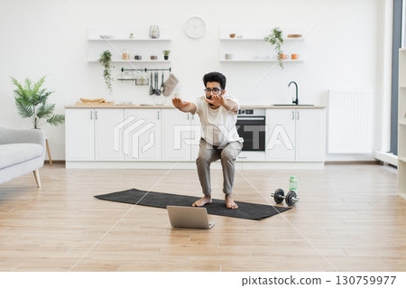 Young adult male exercising indoors on yoga mat following online workout class for fitness 130759977