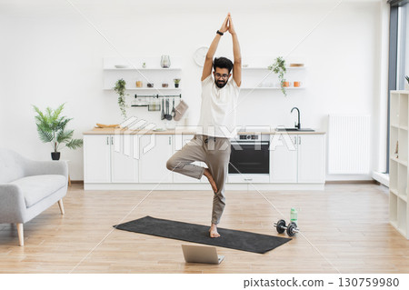 A young adult man performing yoga in home kitchen, recording fitness and wellness content. The serene environment enhances relaxation and video production. A young adult man performing yoga in home kitchen, recording fitness and wellness content. The serene environment enhances relaxation and video production. 130759980