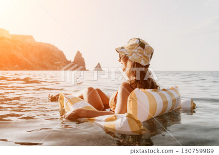 Woman Beach Float Relaxation - A woman relaxes on a yellow and white striped inflatable float in the sea, gazing at the mountains in the distance. 130759989