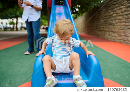 Child Playing on a Slide at the Playground with Adult Supervision Nearby 130763685