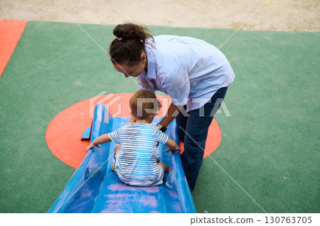 Mother Assisting Toddler on Playground Slide for Fun and Safety 130763705
