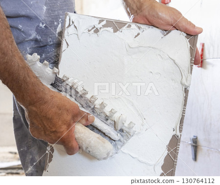 Craftsman spreads tile adhesive with notched trowel on a tile back, closeup. Construction process 130764112