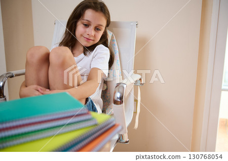 Thoughtful Young Girl Sitting With Colorful Notebooks on a Desk in a Room Thoughtful Young Girl Sitting With Colorful Notebooks on a Desk in a Room 130768054