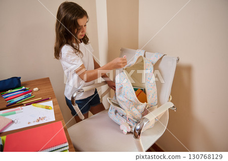 Young Girl Organizing School Supplies Into Backpack on Chair Young Girl Organizing School Supplies Into Backpack on Chair 130768129