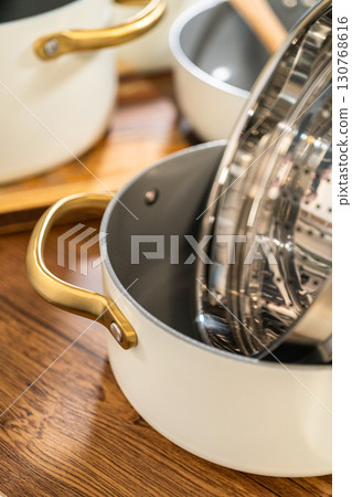 A collection of white enamel cookware with gold handles and metal inserts sits on a wooden kitchen table. The scene features natural lighting and warm tones. A collection of white enamel cookware with gold handles and metal inserts sits on a wooden kitchen table. The scene features natural lighting and warm tones. 130768616