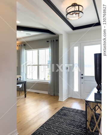 Spacious entryway featuring a coffered ceiling, patterned black rug, and wide windows with curtains. Light walls and natural wood flooring give the space an inviting look. 130768627