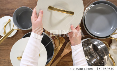 One hand lifts a white lid from a gray saucepan in a tidy kitchen setting. Additional pots, utensils, and a colander fill the rustic workspace. 130768630