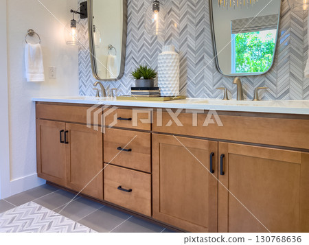 Wide-angle view of a bathroom with dual mirrors, a long wood vanity, and decorative accessories. Herringbone tile adds texture and visual interest to the bright space. Wide-angle view of a bathroom with dual mirrors, a long wood vanity, and decorative accessories. Herringbone tile adds texture and visual interest to the bright space. 130768636