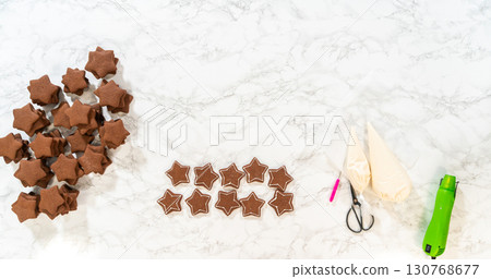 The first step of decorating gingerbread star cookies, showing royal icing tools, including a piping bag filled with white royal icing, placed next to the baked cookies. 130768677