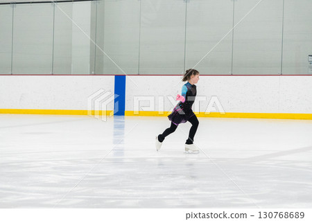 Young girl perfecting her figure skating routine while wearing her competition dress at an indoor ice rink. Young girl perfecting her figure skating routine while wearing her competition dress at an indoor ice rink. 130768689