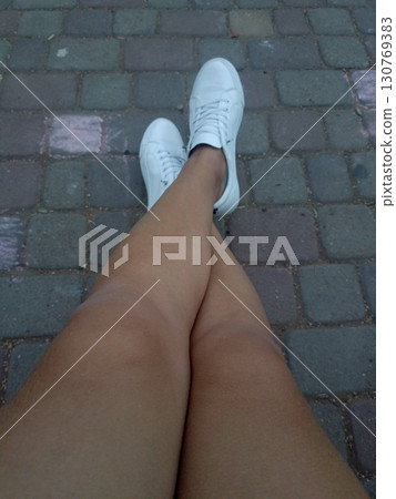 Relaxed person's legs in white sneakers crossed at the ankles on a gray pavement Relaxed person's legs in white sneakers crossed at the ankles on a gray pavement 130769383
