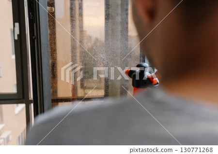 Rear view of janitor male spraying cleaning solution onto large window, preparing to wipe and polish to sparkling shine, ensuring clear and unobstructed view of city outside. Concept of domestic work. Rear view of janitor male spraying cleaning solution onto large window, preparing to wipe and polish to sparkling shine, ensuring clear and unobstructed view of city outside. Concept of domestic work. 130771268