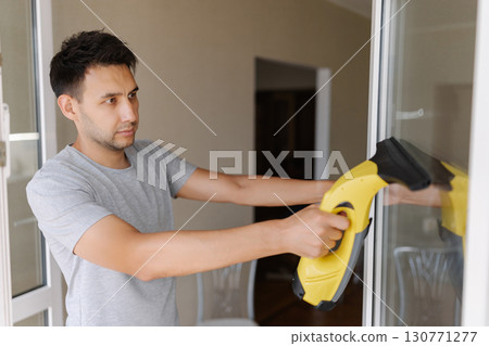 Male worker wiping glass surface using cordless handheld vacuum, working in bright contemporary living space with abundant sunlight streaming inside. Concept of household chores and hygiene. 130771277