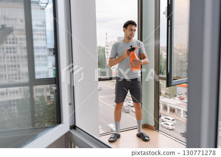 Remote view of homemaker male wearing casual clothes spraying detergent on window in modern apartment, using spray bottle, with blurred city view in background. Concept of cleaning and household. Remote view of homemaker male wearing casual clothes spraying detergent on window in modern apartment, using spray bottle, with blurred city view in background. Concept of cleaning and household. 130771278