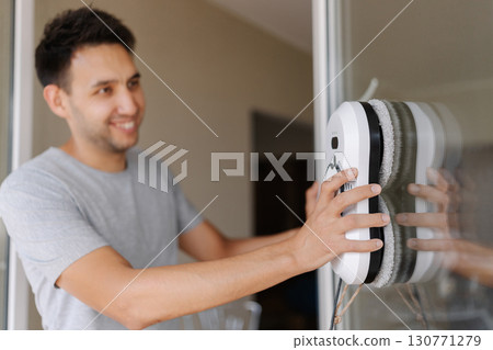 Selective focus shot of smiling young man installing robotic vacuum cleaner on window, preparing to activate device for efficient cleaning session, ensuring spotless and sparkling interior environment Selective focus shot of smiling young man installing robotic vacuum cleaner on window, preparing to activate device for efficient cleaning session, ensuring spotless and sparkling interior environment 130771279