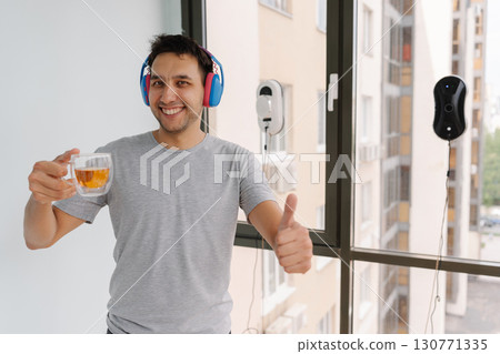 Portrait of happy young man wearing headphones giving thumbs up looking at camera while enjoying tea as automatic window cleaning robot works in background. Concept of cleaning with smart devices. 130771335