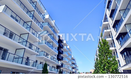 Modern apartment building with balconies and greenery. Beautiful view of real estate buildings in summer. Walking area with trees and grass. Modern residential buildings in the public green area.  130772516