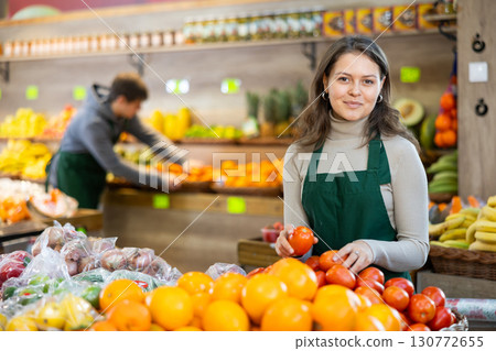 Young female seller holding tomatoes standing by counter in grocery market 130772655