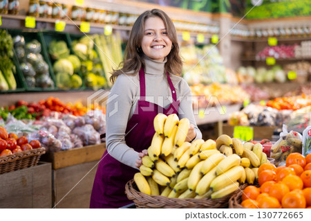 Young female seller holding bananas standing by counter in grocery market 130772665