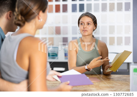 Female salesperson in paint and varnish department is assisting couple customers by showcasing product samples and providing guidance through product catalog at counter 130772685
