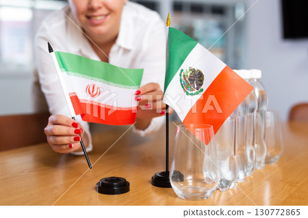 A woman in a white blouse with friendly smile arrangs flags of Mexico and Iran for meeting. 130772865
